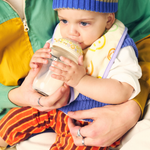Baby drinking from a bottle held by an adult, wearing a colorful outfit and blue hat.