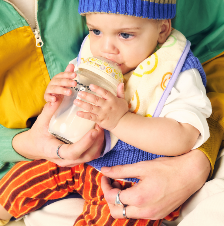 Baby drinking from a bottle held by an adult, wearing a colorful outfit and blue hat.